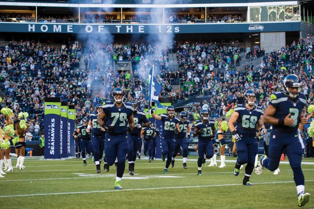 A football team charges onto the field, surrounded by cheerleaders and a roaring crowd in a packed stadium. Smoke fills the air, while a large banner overhead reads