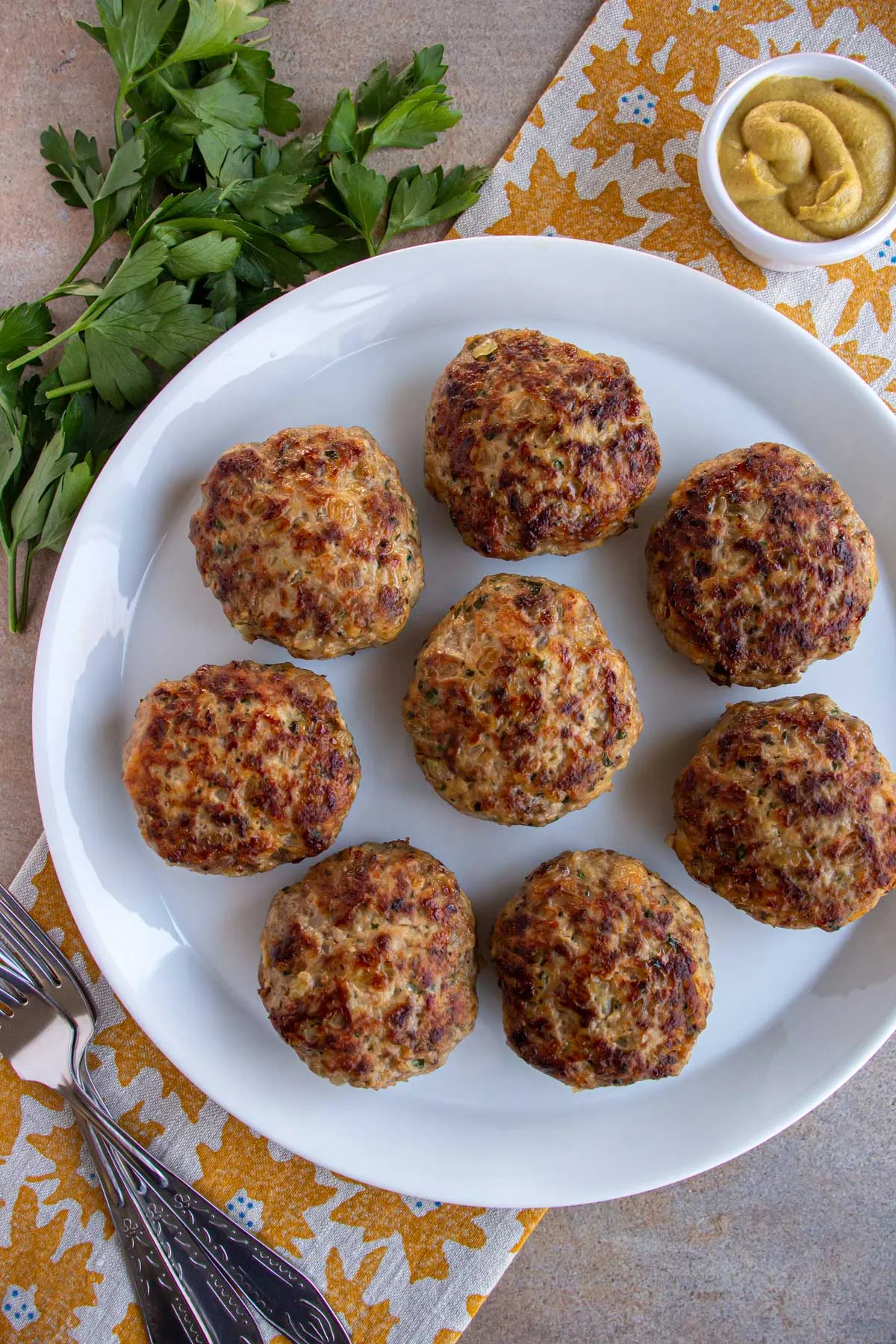 Acht saftige Buletten auf einem weißen Teller, angerichtet auf einem Geschirrtuch mit Sonnenblumenmuster, bereit zum Servieren.