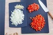 Alle vorbereiteten Zutaten für den Djuvec Reis: fein gewürfelte Zwiebeln, Knoblauch, Paprika und Tomaten auf einem Schneidebrett.