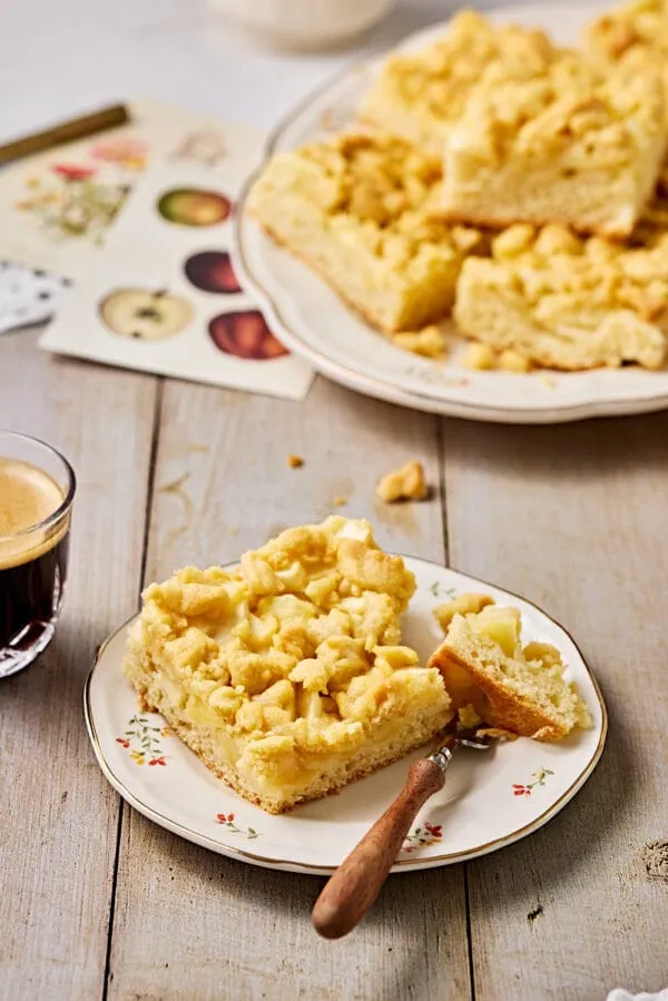Apfelkuchen mit Hefeteig und Streusel auf einer Servierplatte mit Kuchentellern und einem Kaffee im Vordergrund.