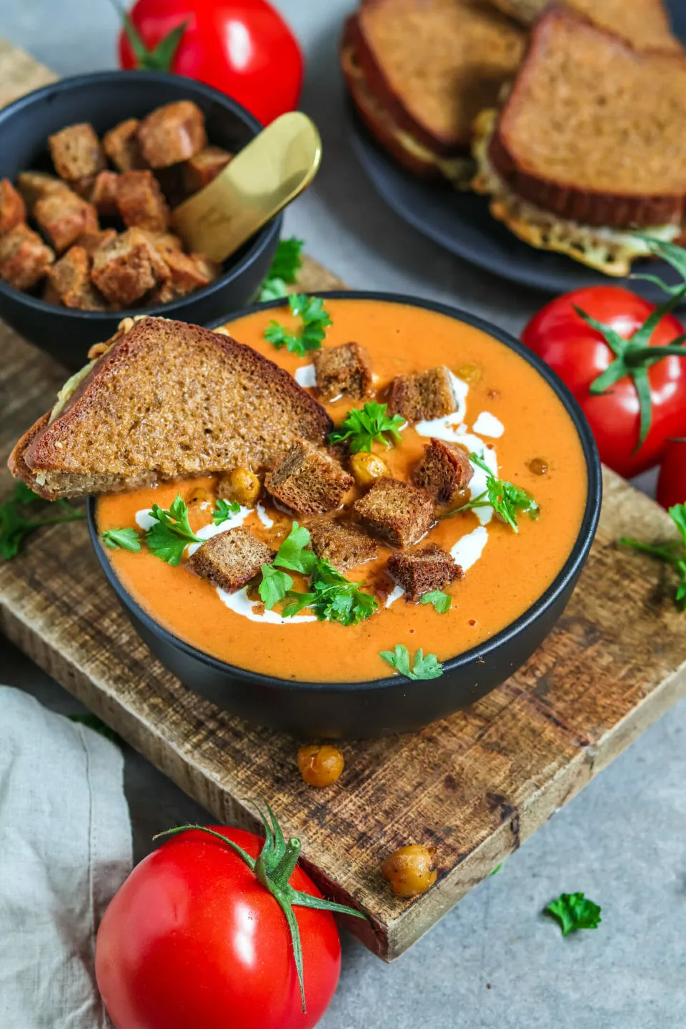 Appetitliche vegane Tomatensuppe, cremig-rot, umgeben von hausgemachten Croutons, knusprigen Kichererbsen und einem veganen Käse-Sandwich als Beilage.