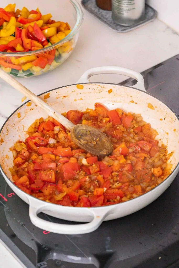 Auf einem Herd steht eine weiße Pfanne mit gehackten Tomaten und Zwiebeln, die mit einem Holzlöffel angebraten werden. Im Hintergrund befindet sich eine Glasschüssel mit einer Auswahl an gehackten Paprikaschoten.