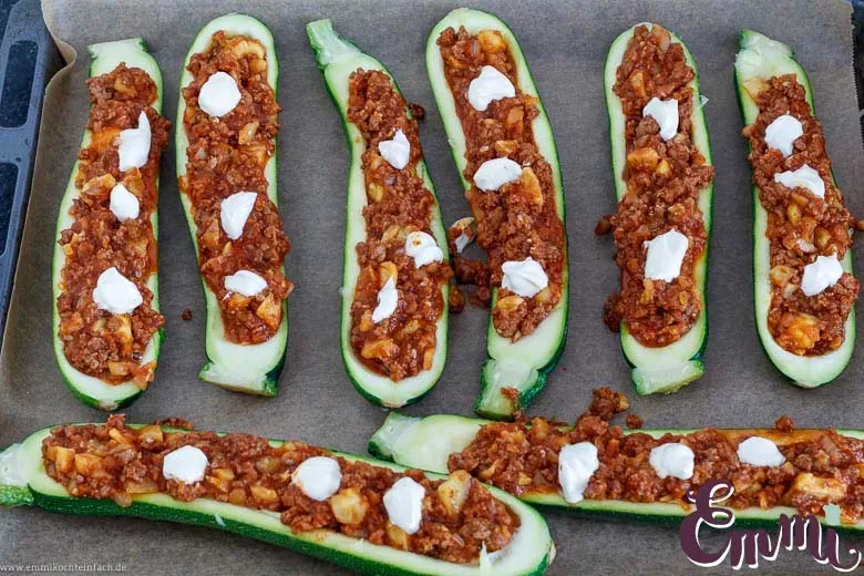 Ausgehöhlte Zucchinihälften, gefüllt mit der herzhaften Hackfleisch-Tomaten-Füllung und Klecksen Crème fraîche, bereit für den Ofen