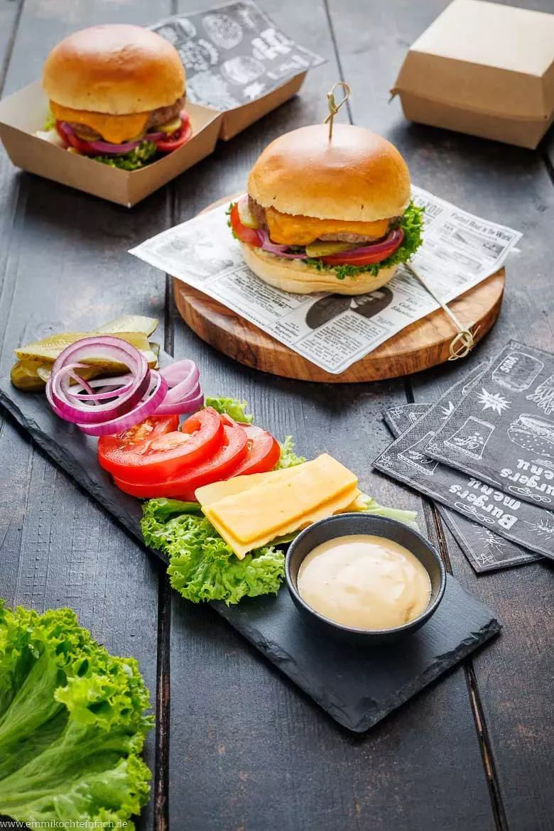 Ausgewählte Zutaten auf einem Holzbrett angerichtet: Burgerbrötchen, Rinderhackfleisch, Salat, Tomaten, Zwiebeln, Käse und Dips.
