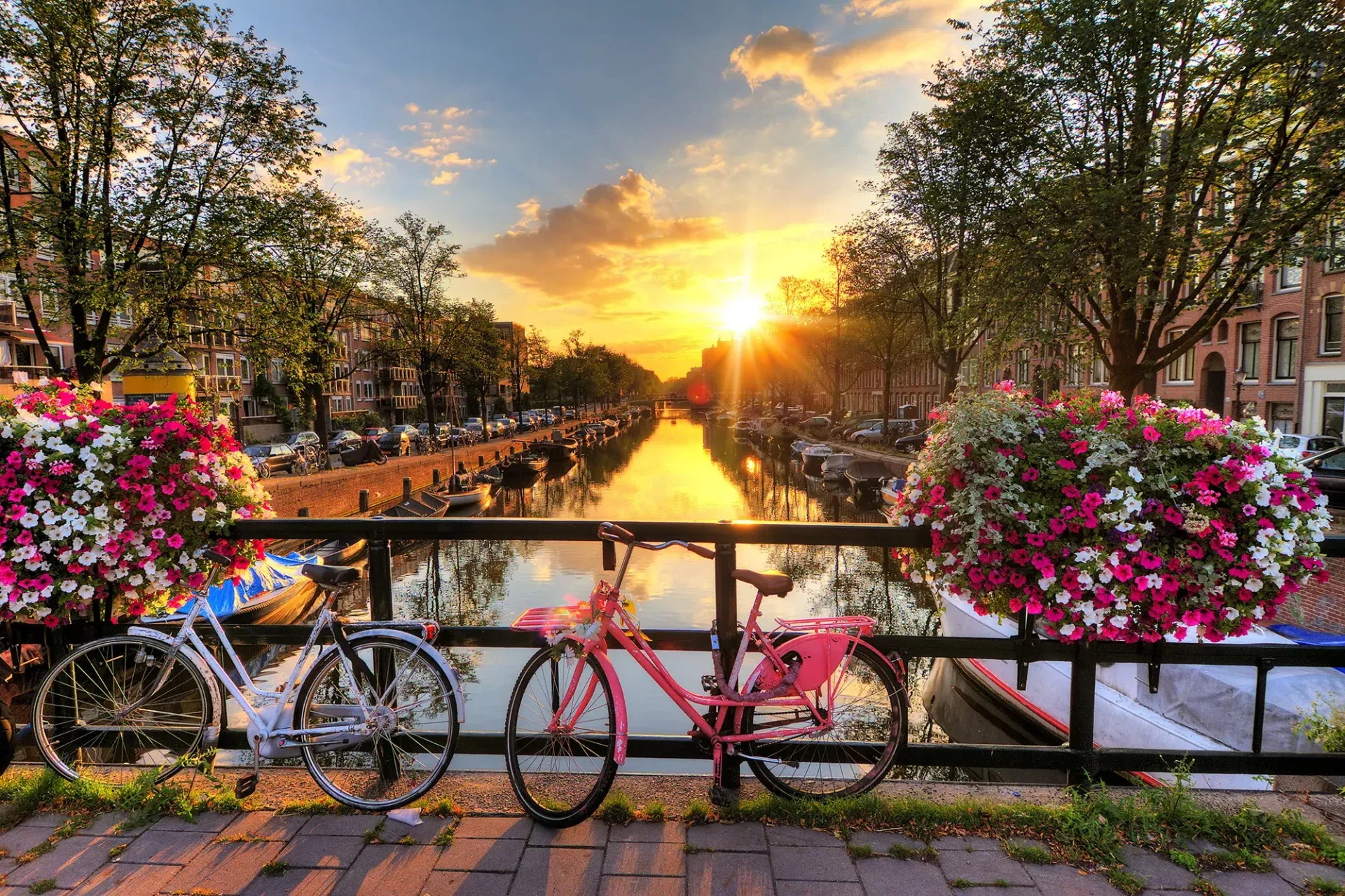 Blick von einer Brücke auf die Gracht bei Sonnenuntergang in Amsterdam, mit angelegten Booten und bunten Fahrrädern.