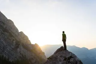 Boulderer steht auf Felsblock, die Sonne geht hinter einem Bergkamm unter