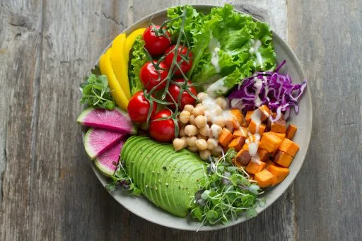 Bunte Buddha-Bowl mit Avocado, Kichererbsen, Kirschtomaten, Süßkartoffeln, Salat, Rotkohl, Mango und Radieschen auf Holztisch