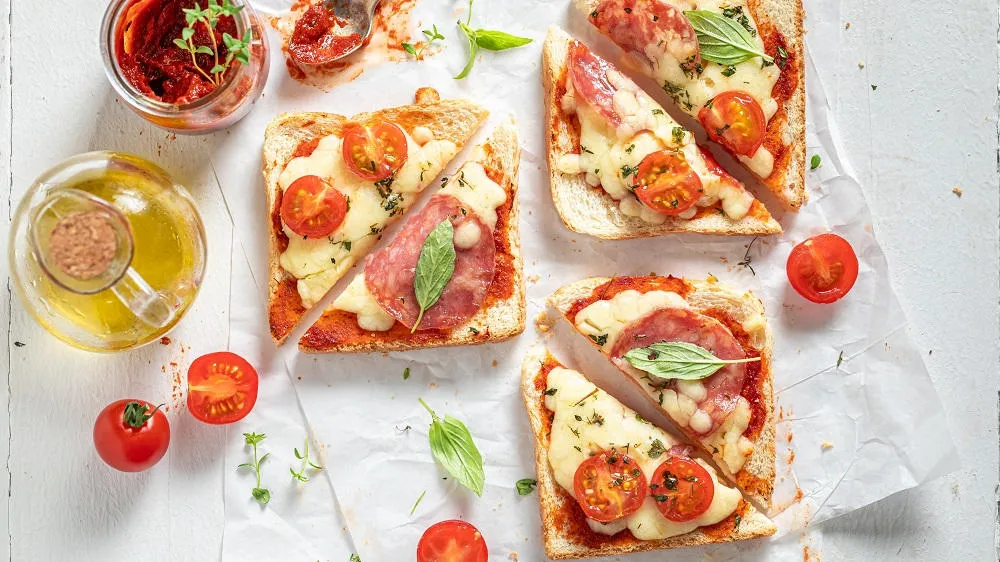 Bunte Pizza-Toasts belegt mit Salami, Pilzen, Oliven und Käse, bereit für den Ofen als unkompliziertes schnelles Abendessen.