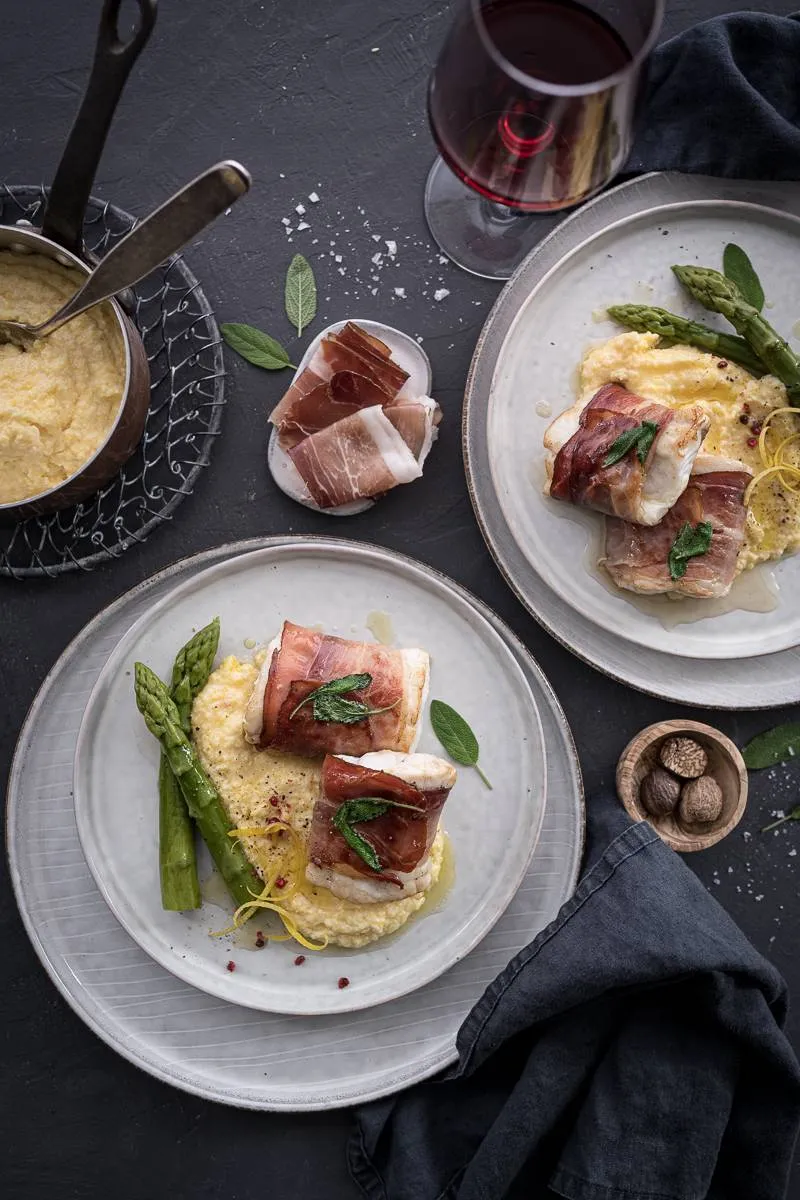 Detailaufnahme eines Tellers mit Zander Saltimbocca, der die Textur des Fisches, die Knusprigkeit des Specks und die Cremigkeit der Polenta zeigt.