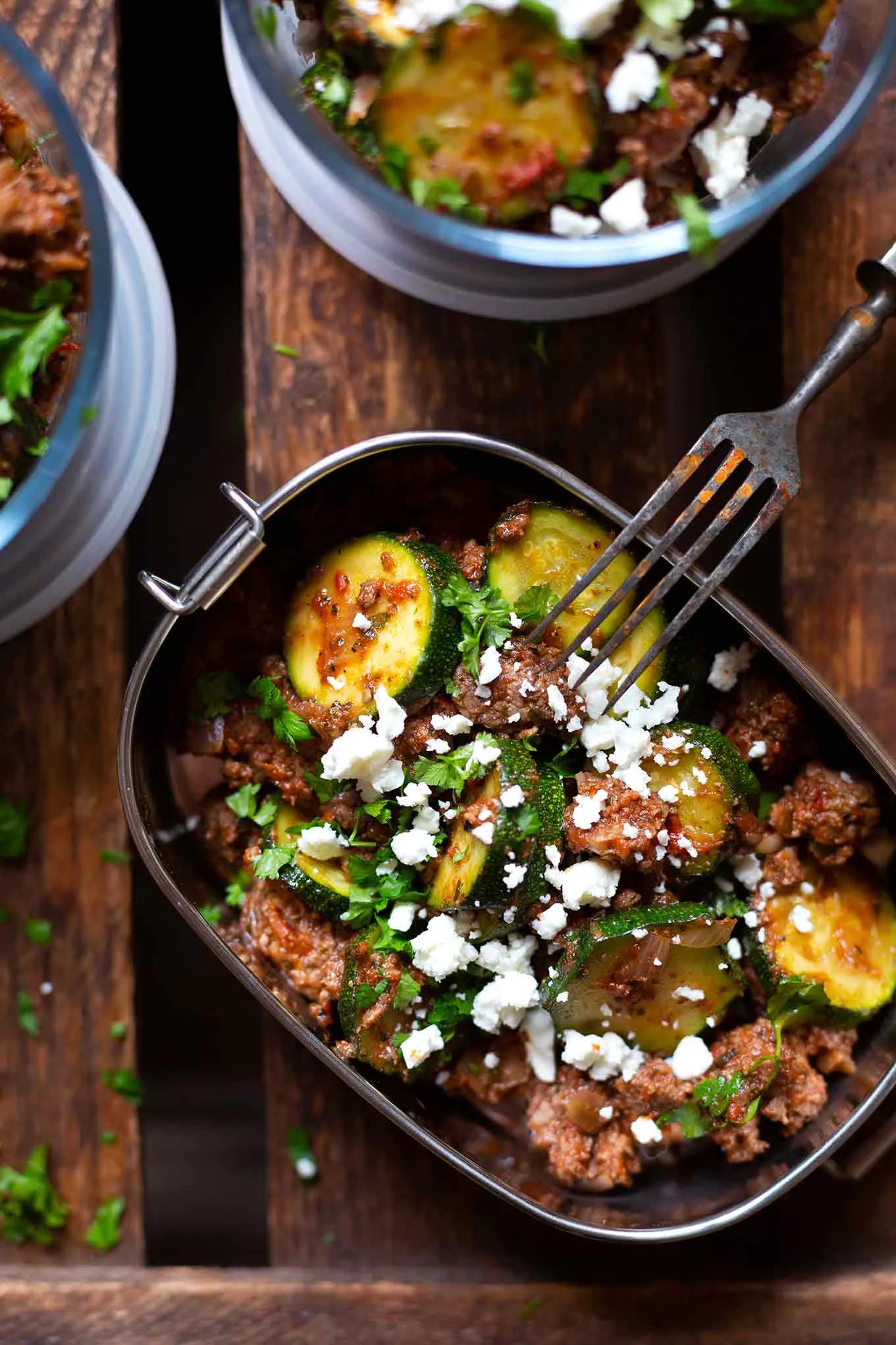 Die fertige Low-Carb-Zucchini-Hackfleisch-Pfanne mit Tomaten und Feta in einer silbernen Meal-Prep-Box. Daneben zwei weitere Meal-Prep-Boxen aus Glas, gefüllt mit der schnellen Zucchini-Hackfleisch-Pfanne. Die Boxen stehen auf einem dunklen Holzuntergrund, und an der silbernen Box lehnt eine Gabel. Das Gericht eignet sich gut zum Mitnehmen.
