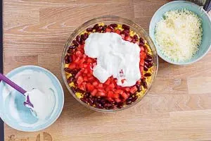 Die letzten Schichten des Taco Salats: cremiger Schmand und geriebener Gouda werden in die Glasschüssel gegeben, bevor die Tortilla-Chips folgen.