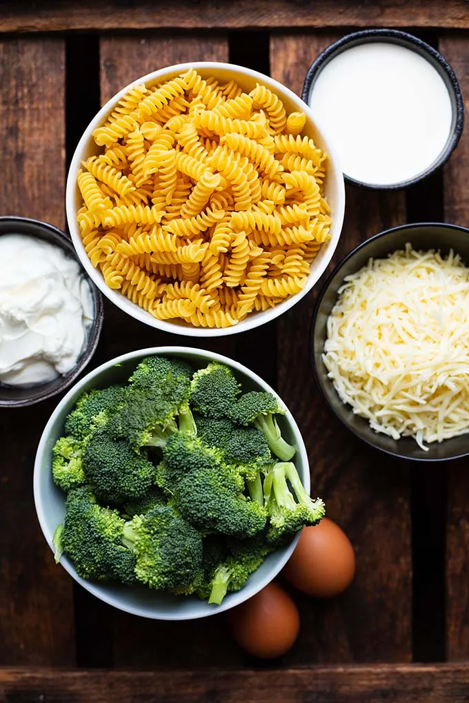 Die Zutaten für den Brokkoli-Nudelauflauf aufgeteilt auf Schüsseln auf dunklem Holzuntergrund. Zu sehen sind, Fusilli-Nudeln, Sahne, geriebener Käse, Brokkoli, Eier und Schmand.