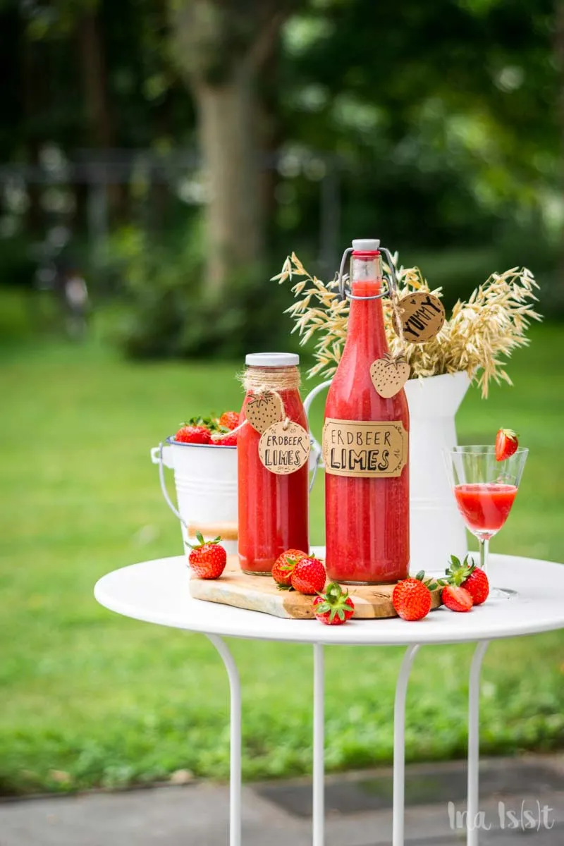 Drei Flaschen frisch zubereiteter Erdbeerlimes auf einem Holztisch, umgeben von grünen Blättern