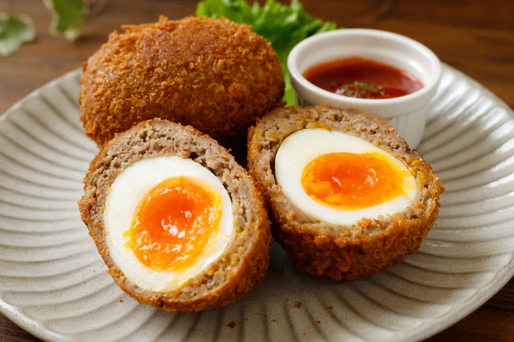 Drei goldbraun panierte Scotch Eggs mit Wurstbrät und hartgekochtem Ei, serviert auf einem Holztisch, bereit zum Verzehr.