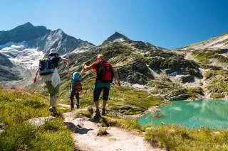 Drei Wanderer, darunter ein Kind, halten sich an den Händen und gehen auf einem Bergpfad entlang eines türkisfarbenen Sees mit schneebedeckten Gipfeln im Hintergrund.