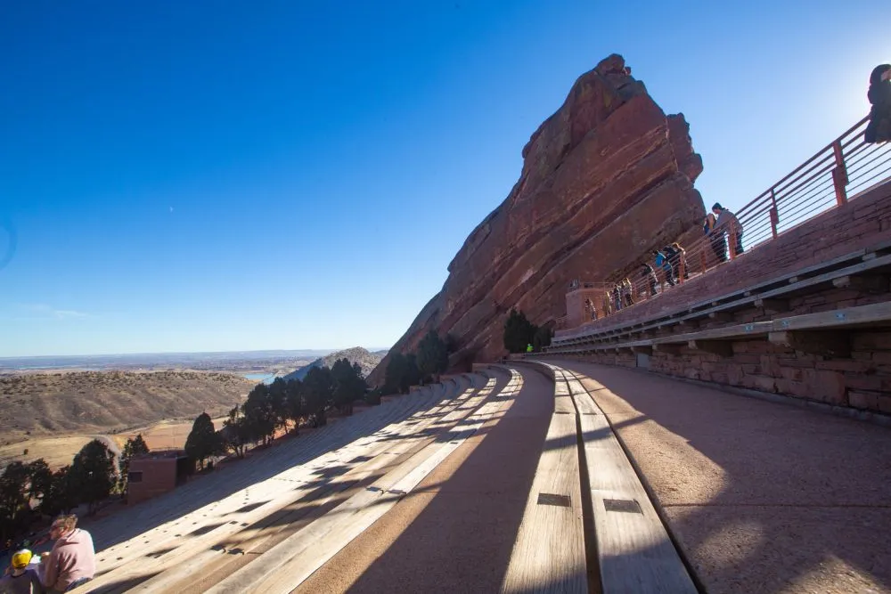 Ein Amphitheater im Freien mit geschwungenen Holzsitzen und einer beeindruckenden roten Felsformation im Hintergrund unter einem klaren Himmel in Colorado. Menschen spazieren am oberen Rand entlang und genießen den weiten Blick auf das Tal.