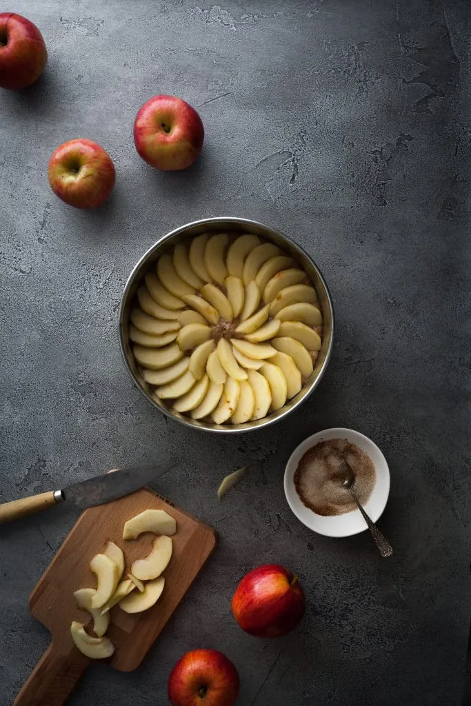 Ein Apfelkuchen in einer Form, daneben eine Schüssel mit Zimtzucker und Apfelspalten auf einem Schneidebrett.