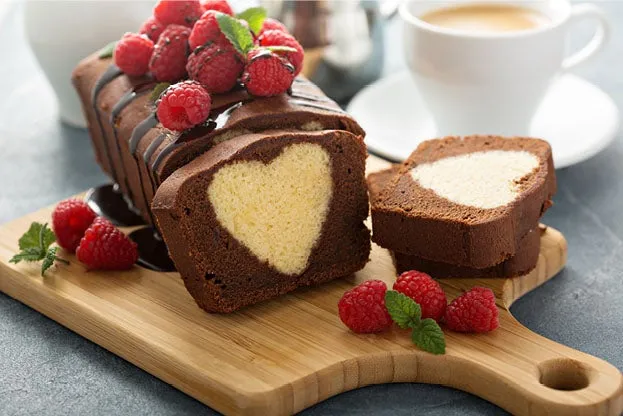 Ein aufgeschnittener Schokoladenkuchen mit hellem Herzmuster im Inneren, garniert mit Himbeeren und Minze, auf einem Holzbrett, neben einer Tasse Kaffee.