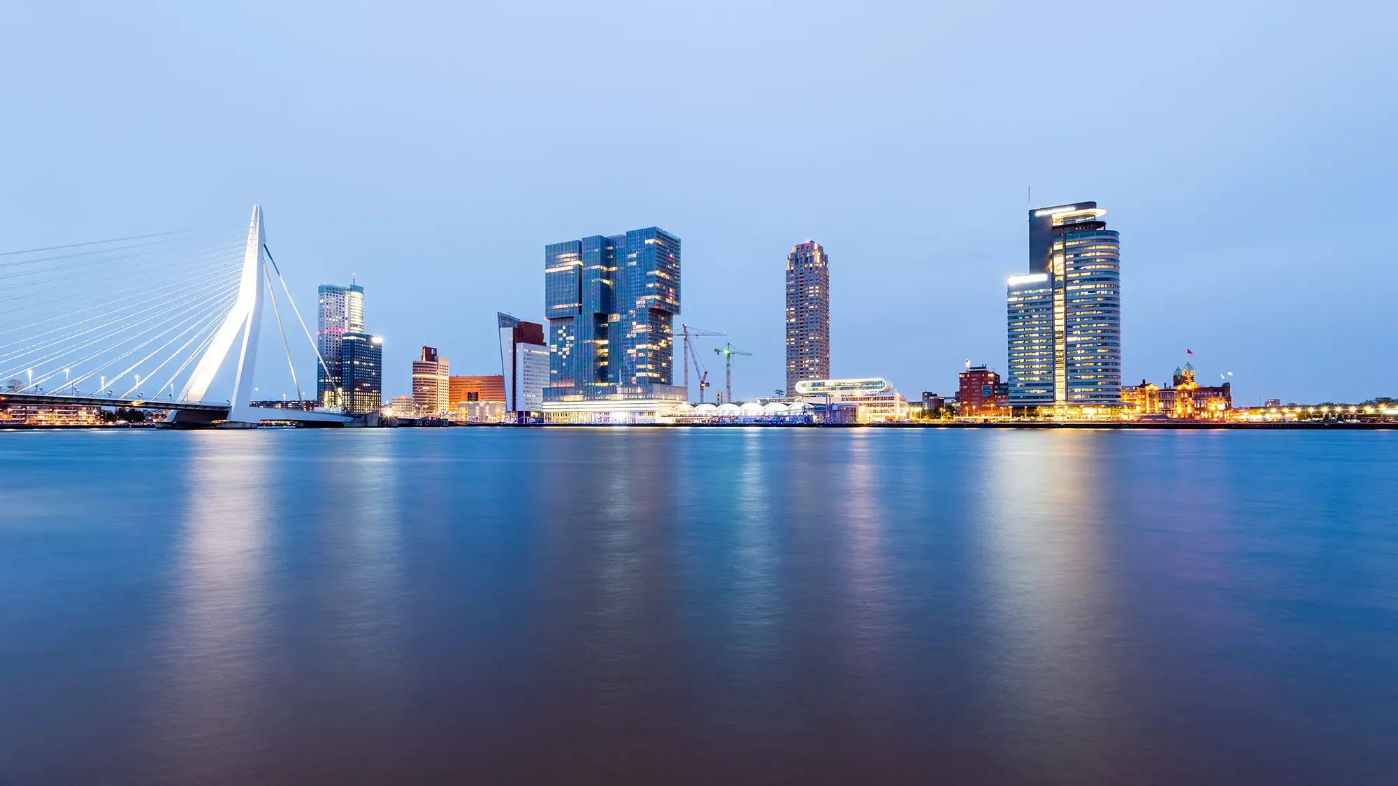 Ein Blick auf die Erasmusbrücke und die beleuchtete Skyline von Rotterdam am Abend mit Reflexionen im Wasser.