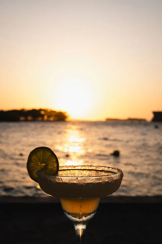 Ein Cocktail im Vordergrund, im Hintergrund ein Sonnenuntergang über dem Meer