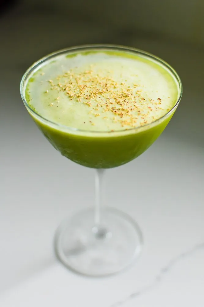 Ein cremiger grüner Matcha-Martini mit Gin in einem Coupé-Glas, garniert mit Muskatnuss auf einer Theke.