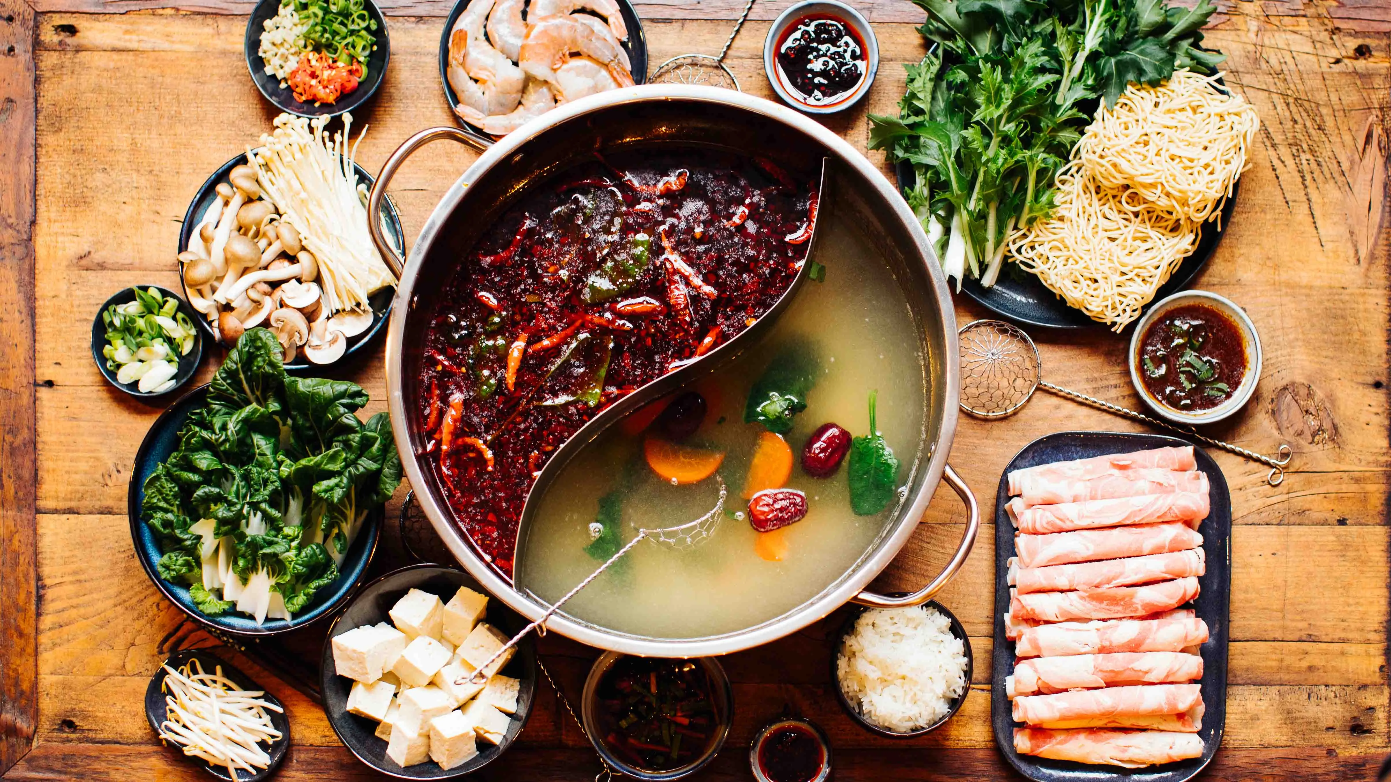 Ein dampfender chinesischer Hot Pot im Szechuan-Stil, umgeben von frischen Zutaten wie Fleisch, Gemüse und Nudeln, für ein geselliges Essen