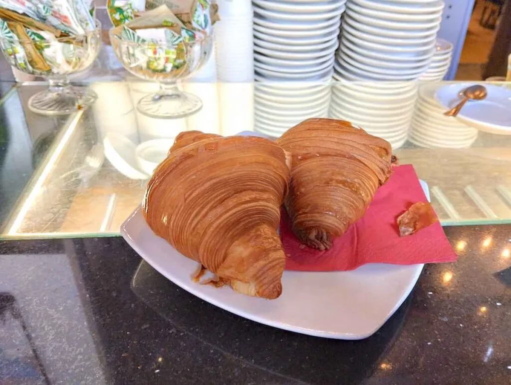 Ein detaillierter Blick auf zwei Croissants mit sichtbar geschichteten, goldenen Teig auf einem weißen Teller, der auf einer gläsernen Arbeitsplatte mit gestapelten weißen Tellern und Glasbehältern mit verpackten Süßigkeiten im Hintergrund ruht, alles unter dem sanften Schein der Deckenbeleuchtung.