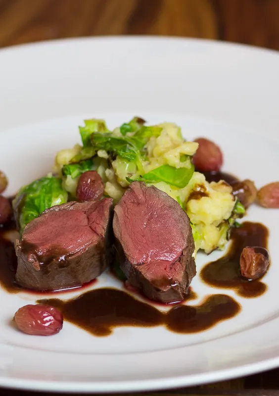 Ein festlich angerichteter Rehrücken sous vide mit Kartoffelstampf, Rosenkohl, Haselnüssen und karamellisierten Trauben, perfekt für ein deutsches Herbstmenü.