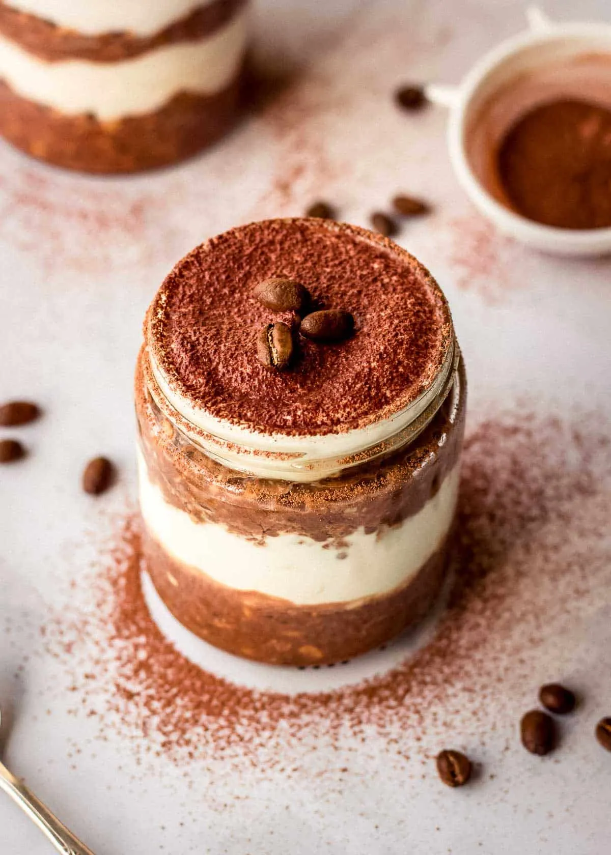 Ein Glasgefäß mit geschichteten Tiramisu Overnight Oats, dekoriert mit Kakaopulver und Kaffeebohnen.