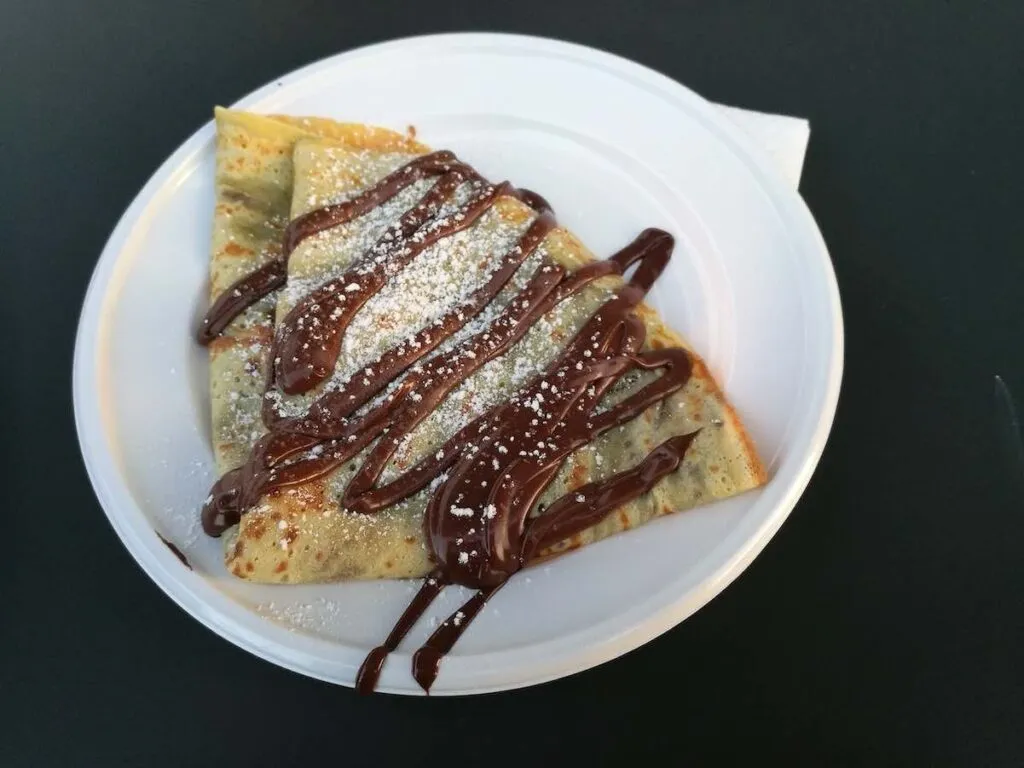 Ein goldbrauner süßer Crêpe, der auf einem weißen Einwegteller gefaltet ist, mit großzügigen Mengen an Schokoladensauce und einer Prise Puderzucker beträufelt, auf einer dunklen Tischplatte serviert.