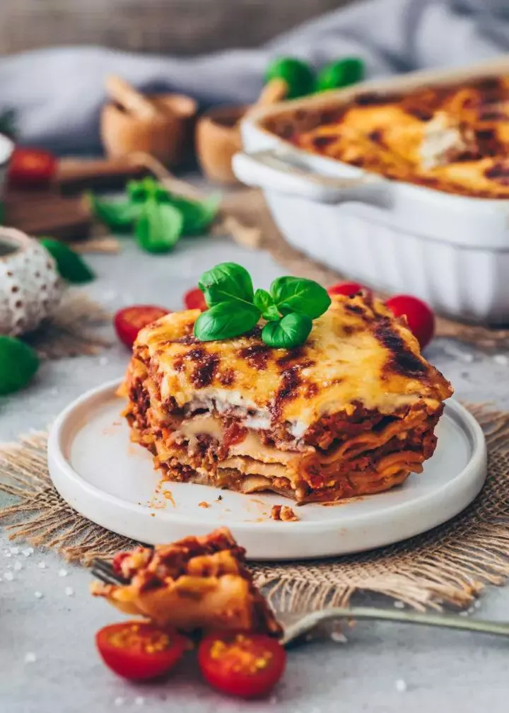 Ein großzügiges Stück veganer Bolognese-Lasagne al Forno auf einem Teller, mit Basilikum garniert und im Anschnitt gut sichtbar.