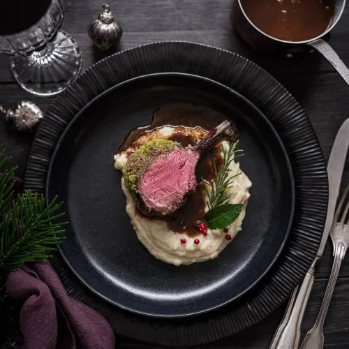 Ein horizontales Rechteckbild, das ein Lammkarree zeigt, das auf einem weißen Teller mit Beilagen serviert wird und mit frischen Kräutern und Gewürzen dekoriert ist.