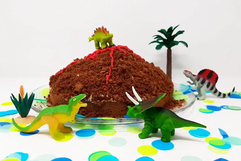 Ein kreativ dekorierter Vulkankuchen mit Schoko-Haselnussboden, cremiger Quark-Sahne-Füllung und Bananen, überzogen mit roter "Lava" aus Cakemelts und kleinen Dinosaurier-Figuren, ideal für einen Kindergeburtstag oder die festliche Kaffeetafel.