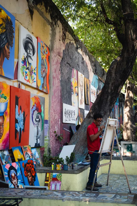 Ein Künstler malt ein Bild auf einer Staffelei unter einem Baum, um ihn herum sind farbenfrohe Porträts und Kunstwerke an einer Mauer aufgehängt