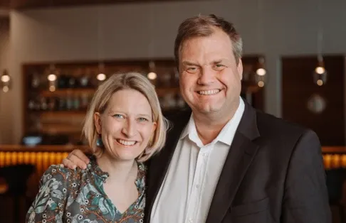 Ein lächelndes Paar steht zusammen in einem warm beleuchteten Raum mit Regalen und Lichtern im Hintergrund.