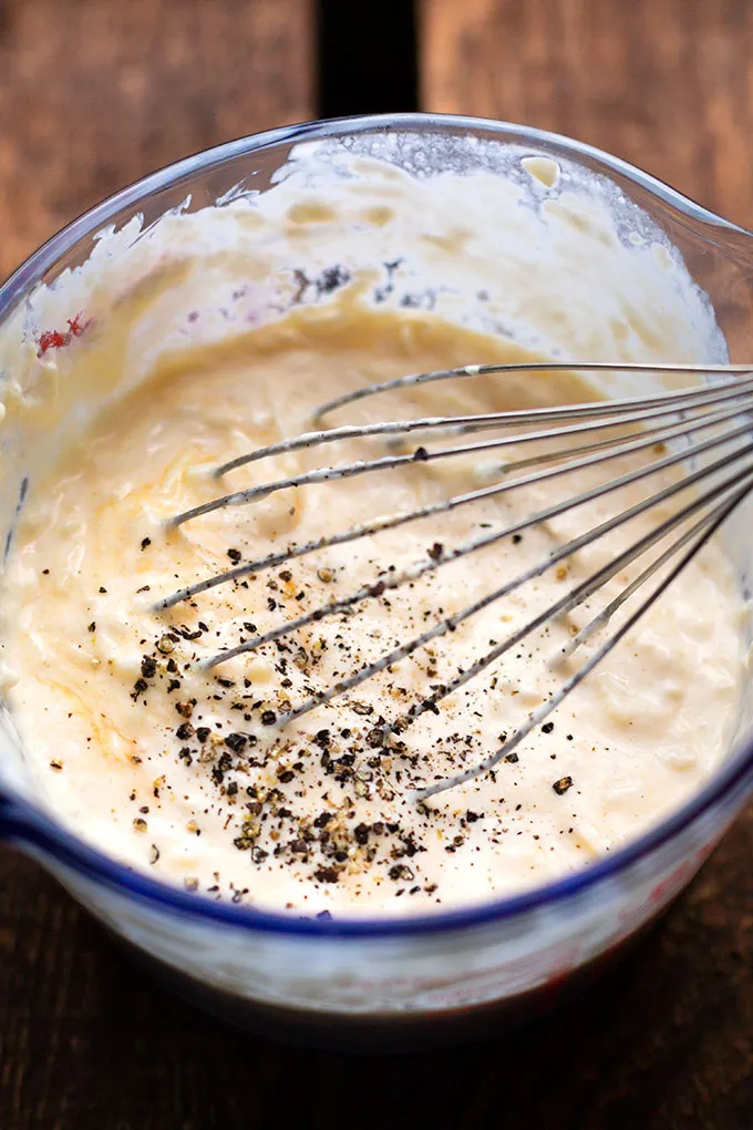 Ein Messbecher aus Glas mit der Soße für den Brokkoli-Auflauf mit Nudeln. Verrührt mit einem Schneebesen und Pfeffer oben drauf.