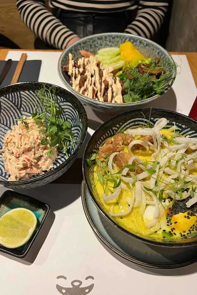 Ein modernes asiatisches Fusionsgericht von Soi Fusion in Zagreb, mit einer Nudelschale mit leuchtend gelbem Curry und knusprigen Wan-Tans, dazu grünes Gemüse und Krabbensalat, geteilt an einem Tisch.