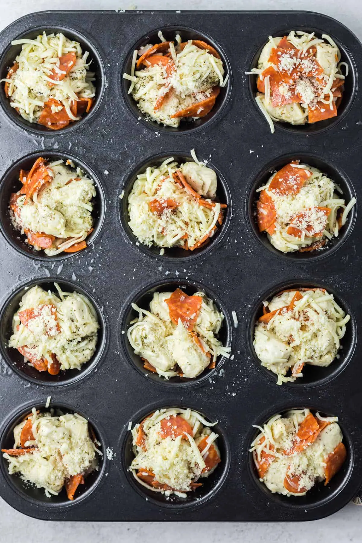 Ein Muffinblech mit zwölf Mulden, gefüllt mit der Pizza Muffin Mischung und mit Parmesan bestreut.