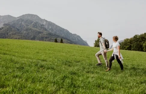 Ein Paar wandert auf einer saftig grünen Wiese mit Bergen im Hintergrund und fängt die natürliche Schönheit in der Nähe des Hotel Klosterhof ein.