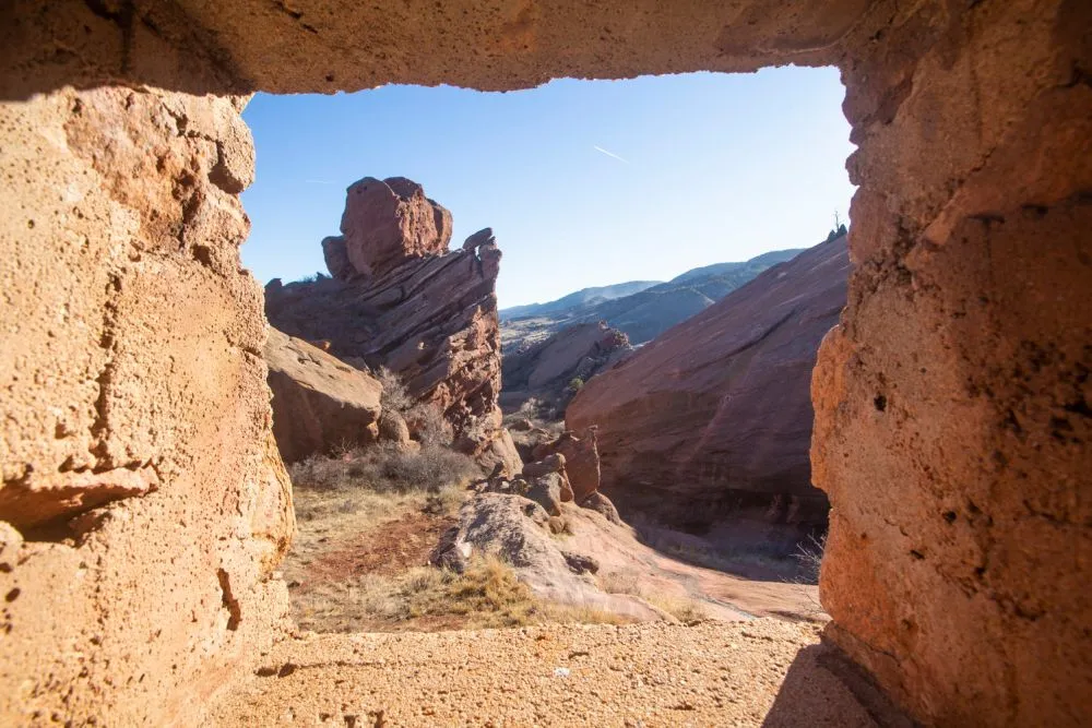Ein Panoramablick durch einen Felsbogen im Red Rocks Park in Colorado, der die zerklüfteten roten Felsformationen und den strahlend blauen Himmel einfängt, wobei das Sonnenlicht dramatische Schatten auf die Landschaft wirft.