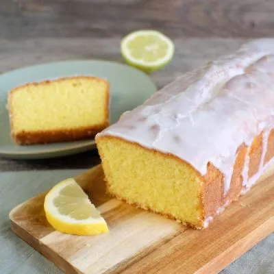 Ein perfekt gebackener glutenfreier Zitronenkuchen mit glänzendem Zitronenguss, aufgeschnitten auf einem Holzbrett, daneben frische Zitronenscheiben und ein Stück auf einem Teller. Er veranschaulicht die Saftigkeit des Kuchens und die Frische der Zutaten.