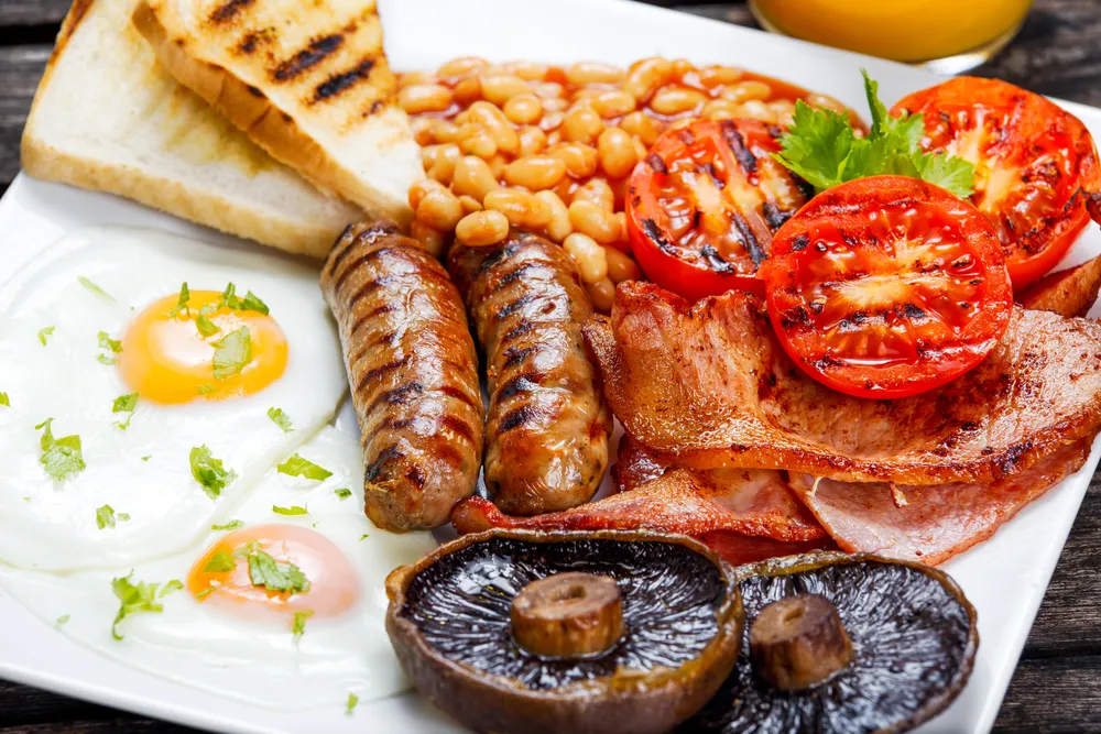 Ein reichhaltiges Full English Breakfast mit Spiegelei, Speck, Würstchen, gegrillter Tomate, Pilzen, Baked Beans und Toast auf einem weißen Teller.
