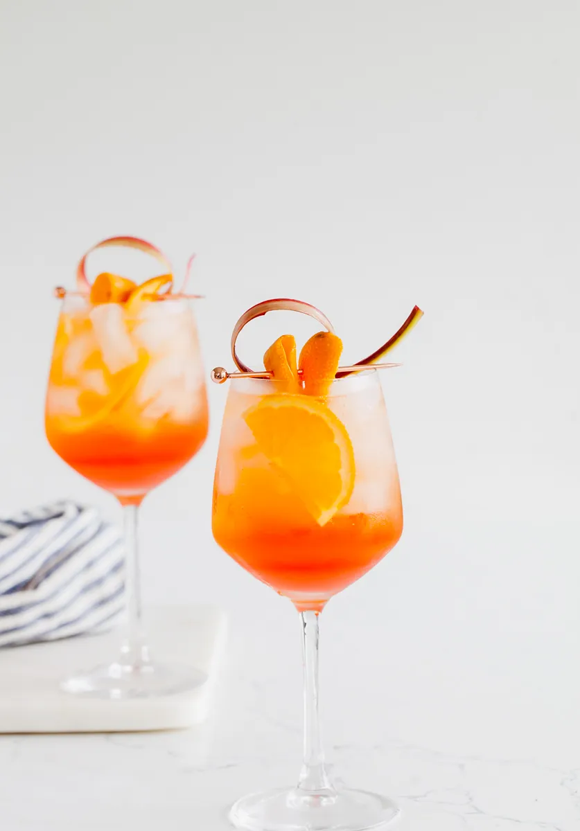 Ein Rhabarber Aperol Spritz mit Rhabarberstreifen und Orangenzeste in einem stilvollen Weinglas.