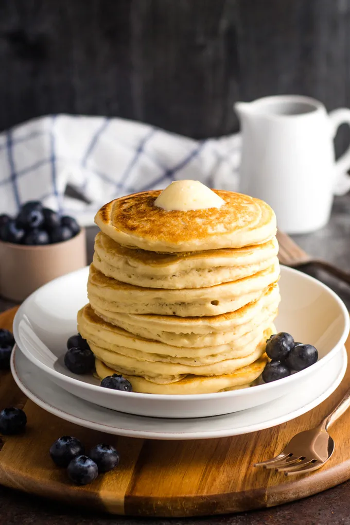 Ein Stapel fluffiger veganer Pancakes, beträufelt mit Ahornsirup und umgeben von Blaubeeren.