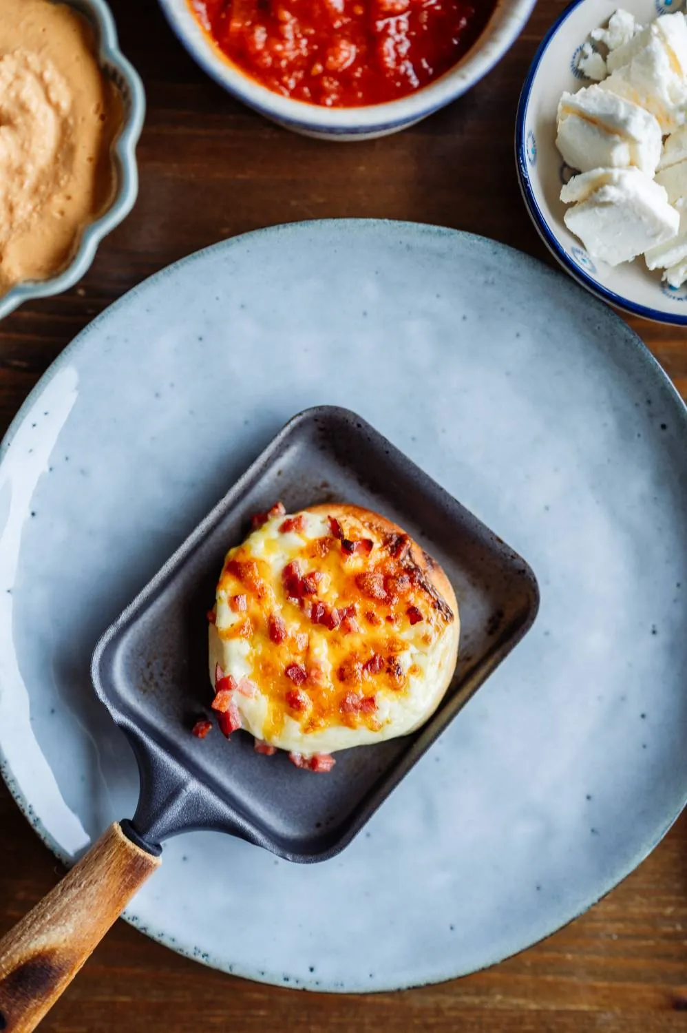 Ein Stück Camembert schmilzt in einem Raclette-Pfännchen, umgeben von weiteren Zutaten