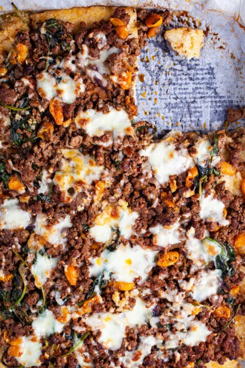 Ein Stück einer Polenta Pizza, belegt mit Hackfleisch, Gemüse und Käse, frisch aus dem Ofen entnommen.