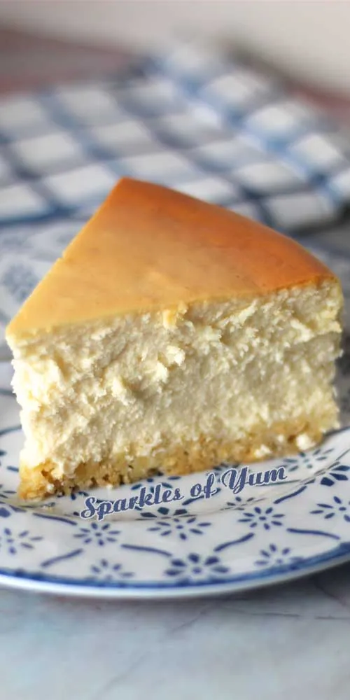 Ein Stück New York Cheesecake auf einem weißen Teller mit blauen Mustern, daneben ein weißes Handtuch mit blauen Streifen im Hintergrund.