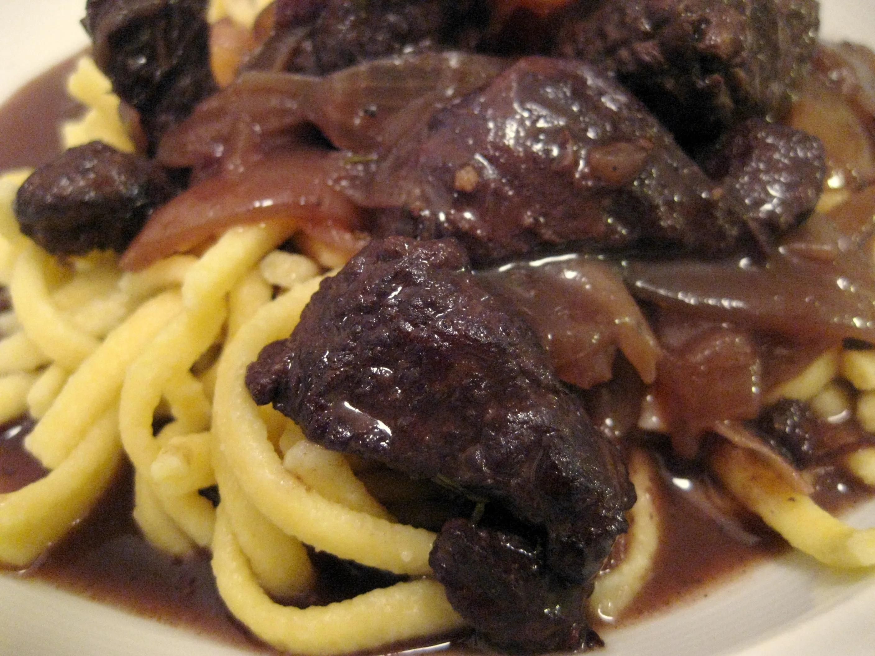 Ein Teller Hirschgulasch mit Spätzle und Preiselbeeren, mit einem Glas Rotwein daneben.