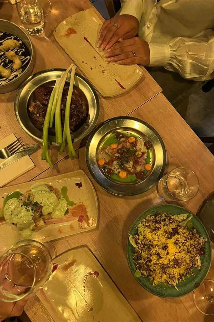 Ein Tisch im Sopal Fun Dining Bar in Zagreb, gedeckt mit verschiedenen Gerichten und Weinflaschen, perfekt für ein geselliges Esserlebnis, wie Schwarzrisotto mit Grieben und geröstetes Lamm.