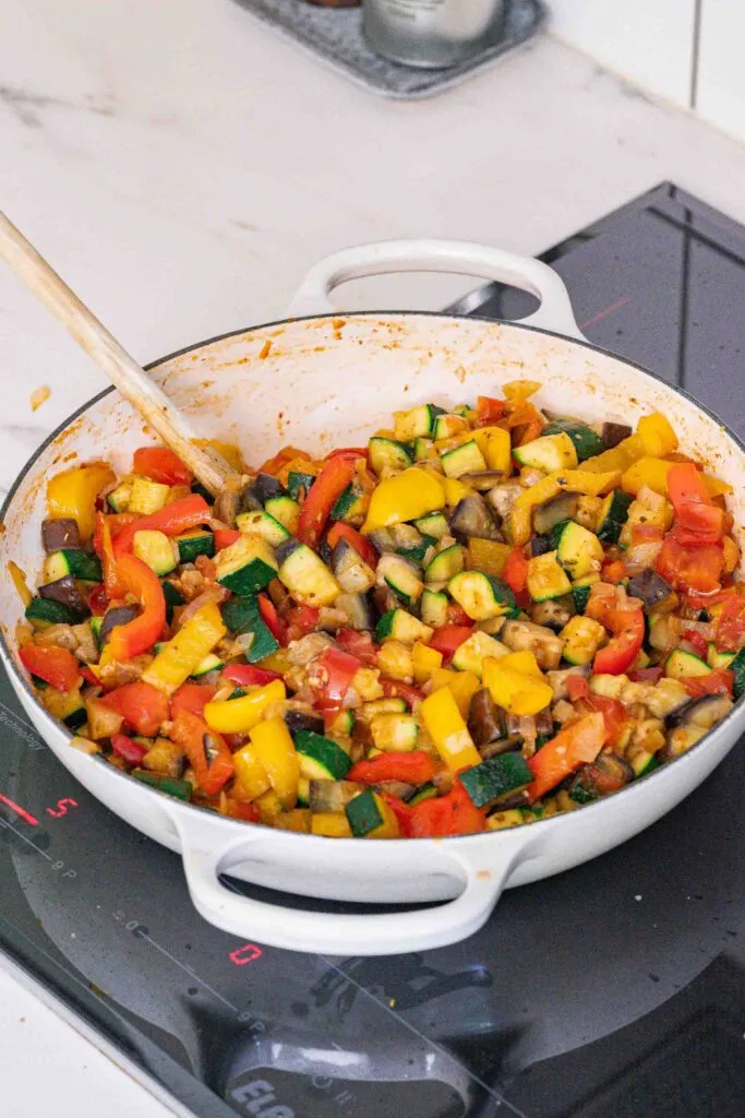 Ein weißer Topf auf einem Herd enthält eine Mischung aus gehacktem Gemüse, einschließlich Zucchini, Paprika und Tomaten, die mit einem Holzlöffel umgerührt werden.
